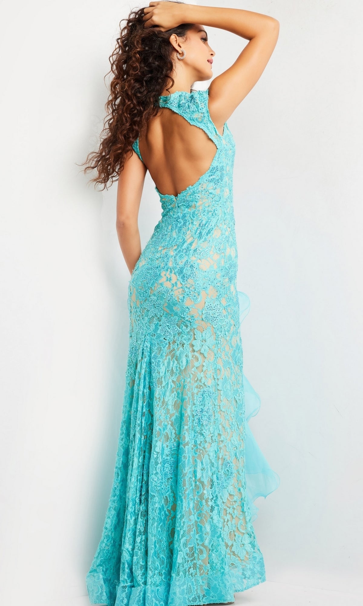A woman stands with one hand on her head, facing away from the camera in a turquoise Jovani Ruffled Long Lace Prom Dress 38668 against a plain light background.